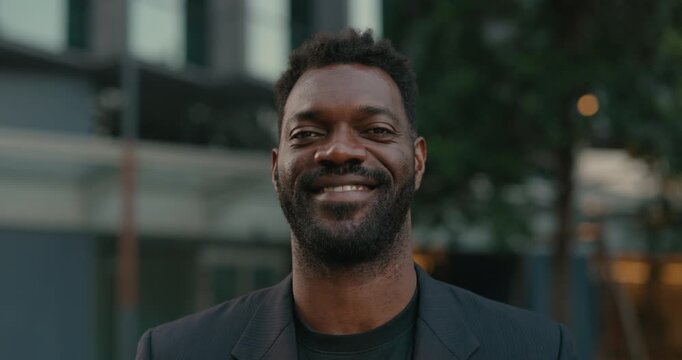 Smiling African American businessman looks at the camera outside a contemporary office, conveying approachable leadership, confidence and professional success.