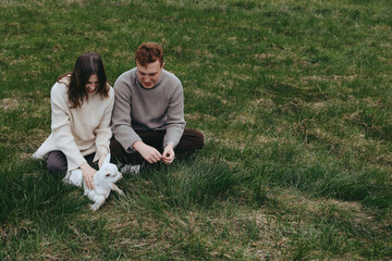 Fototapeta premium A couple sits on a grassy meadow with a goat kid, enjoying a calm outdoor moment, a wholesome lifestyle scene that highlights family, nature, companionship, and simple rural joy.