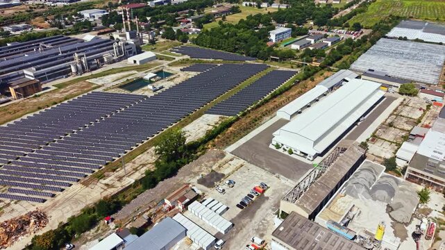 Solar panels are arranged in rows on a flat area beside factories. Buildings and trees are visible in the distance under a clear sky.