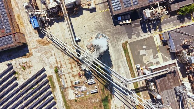 An industrial area is displayed from above with pipes and machines. Solar panels are visible on the ground. Smoke is coming from a section of the site. It is daytime.