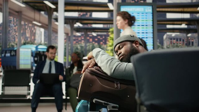 Tired man rests on luggage in a bus station during a long wait for a delayed shuttle, leaning on a travel bag before continuing a trip to another country.