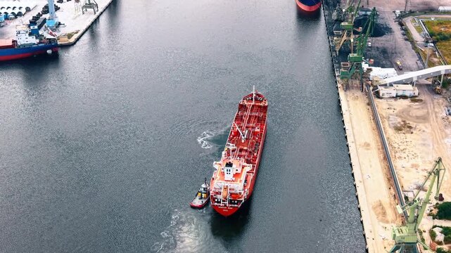 Top down aerial view of red tanker ship with tugboat. Small vessel maneuvers beside large vessel in dark water near harbor docks and tall cranes.