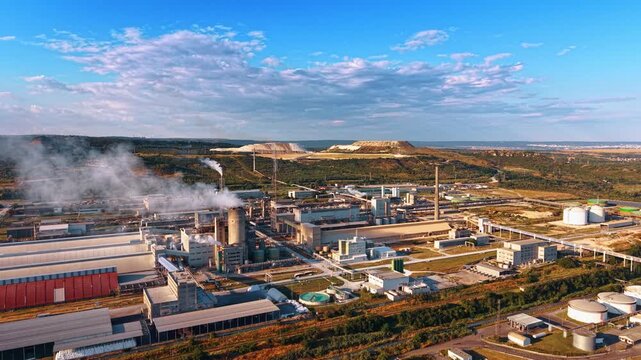 Panoramic aerial view of expansive industrial complex. Multiple factory buildings and pipe systems stretch toward distant hills under blue sky.