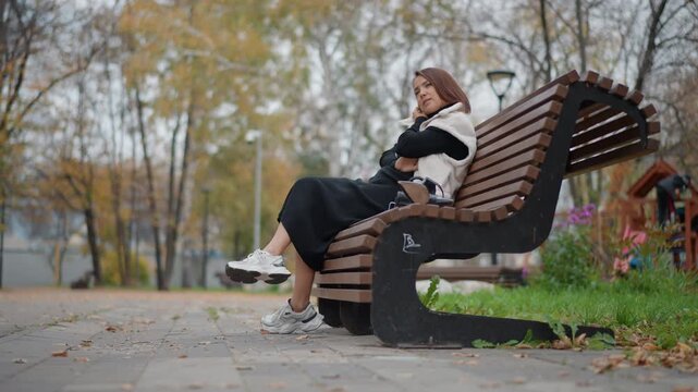 Young woman sits on curved wooden park bench with crossed legs, leaning back and turning upper body to look aside, showing relaxed mood, casual style, soft daylight, and calm outdoor movement