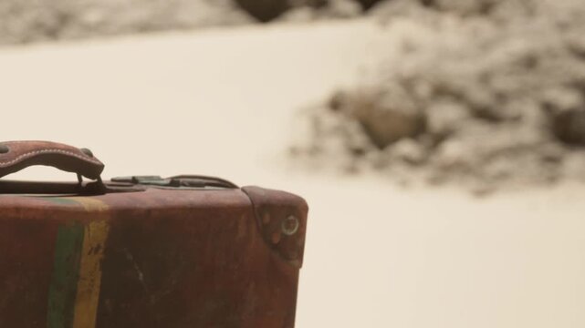 A suitcase sits alone on the sand in a remote area. In the background, there are rocks scattered around the sandy surface. The scene suggests a feeling of solitude and travel.