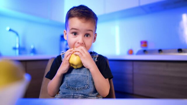 Young boy taking a bite of yellow apple. A child holds fresh fruit with both hands while eating at a kitchen table under blue ambient LED light.