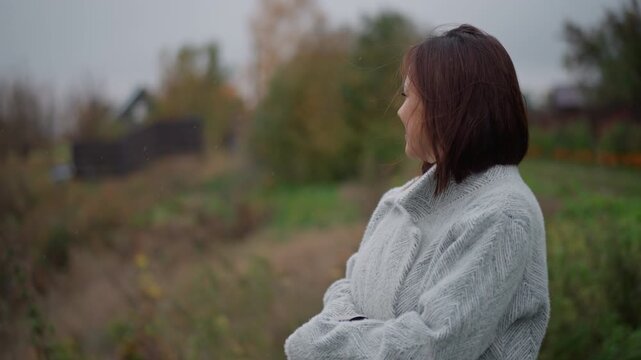 Mature woman stands in side profile on grassy hillside wearing textured knitted shawl with folded arms, steady gaze toward right, gentle breeze moving hair and shawl edges under soft overcast light