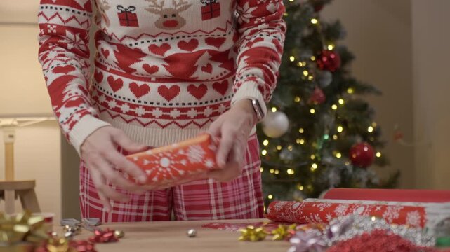 Woman wearing Christmas sweater placing gift into wrapping paper at table with decorations and ribbons. Concept of holiday preparation