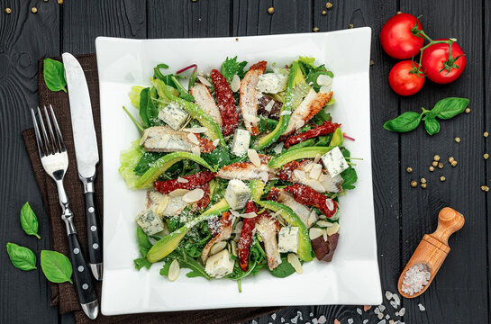 Flat lay salad plate with avocado smoked eel and cheese. Square white dish with arugula avocado slices sun dried tomatoes and cutlery on black wooden surface.