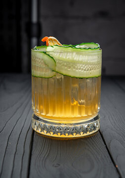 Whiskey glass with cocktail cucumber slice and orange garnish. Yellow cocktail served in textured glass garnished with cucumber slices and dried orange.