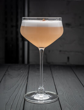 Layered sour cocktail in a stemmed coupe glass. Front view displays a peach drink with thick white foam on a dark wooden surface.