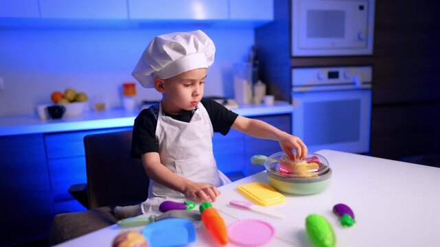 Young boy placing plastic toy food in toy pan. Child wearing chef toque and apron sits at table with toy cooking set.
