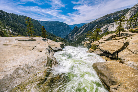 Fast-flowing river cascading over the edge of a granite cliff along the Mist Trail, overlooking a deep green valley in Yosemite National Park.