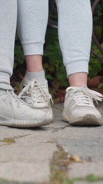 Feet wearing two sets of shoes with one pair new clean. One foot has newer clean sneaker while opposite shoe. Background contains moss or grass growth on paved area where