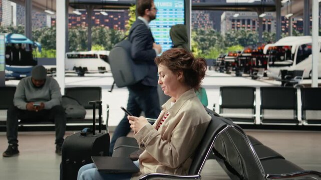 Smiling woman texts on a touchscreen smartphone in a bus station while waiting for an intercity coach connection, chatting with friends online on her mobile phone during the wait before departure.