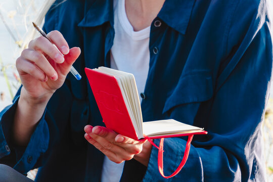 Close up view of hands holding a red notebook and pen while taking notes outdoors