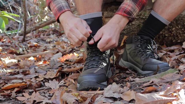 Man in a plaid shirt tying his hiking boots. He is sitting on a log in a forest, surrounded by fallen autumn leaves. Active lifestyle concept. Natural light, earth tones.
