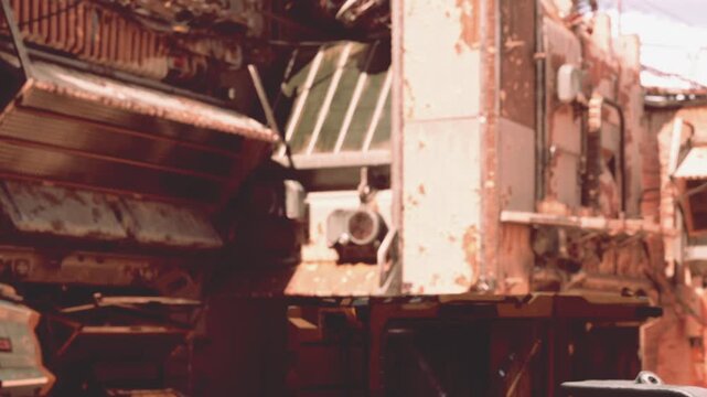 Parts of a broken factory are seen in bright sunlight. Rust covers many surfaces. Tools and machines are scattered around the site. This location is near a busy city square.