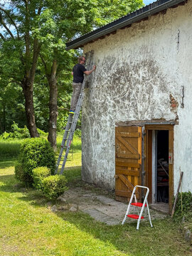 Man on ladder renovating weathered barn wall in garden