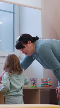 Adult guides child at colorful toy filled display indoors. Child reaches for items at toy display as adult leans. Setting includes window backdrop for woman guiding young