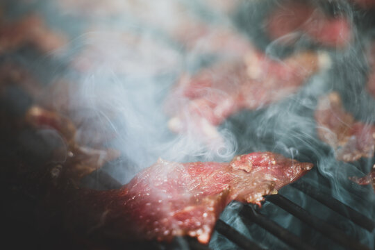 A close‑up of round steak slices cooking on a pellet grill smoker shows rich color and rising smoke, emphasizing the heat and aroma of beef jerky preparation.