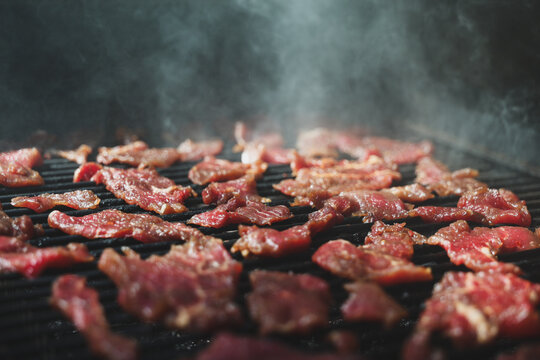 Thin strips of marinated round steak sizzle on a pellet grill smoker, surrounded by rising smoke and glowing heat, illustrating the art of homemade beef jerky.