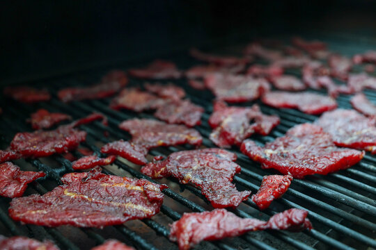 Thin slices of round steak dry slowly on a pellet grill smoker, their deep red color and crisp texture revealing the final stage of homemade beef jerky creation.