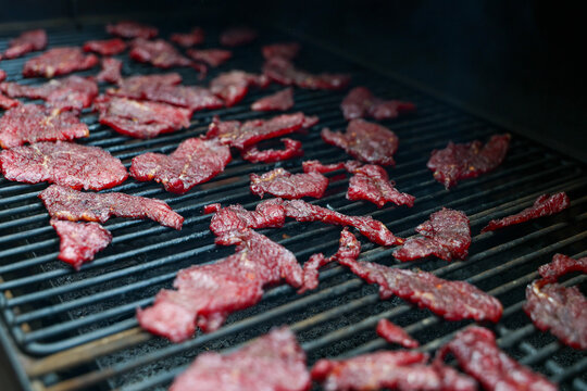 Thin slices of round steak dry slowly on a pellet grill smoker, their deep red color and crisp texture revealing the final stage of homemade beef jerky creation.