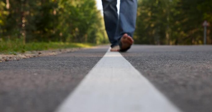 A barefoot pedestrian in blue jeans on the road, one barefoot man walks along the road along the white marking line