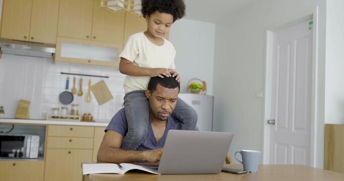 Busy black African American father using a laptop while young child plays on his shoulders, illustrating remote work, parenting challenges, and family life.