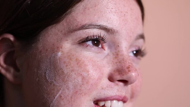 Beautiful girl with freckles applying cream on her face against beige background, closeup