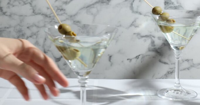 Woman pouring alcohol drink into glass while her friend taking martini cocktail with olives at white table, closeup. Camera moving left
