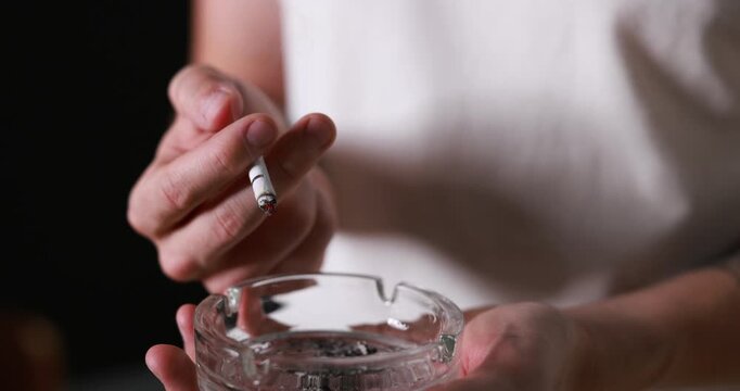 Man flicking ash from cigarette into ashtray indoors, closeup. Camera moving in