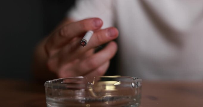 Man flicking ash from cigarette into ashtray at table indoors, closeup