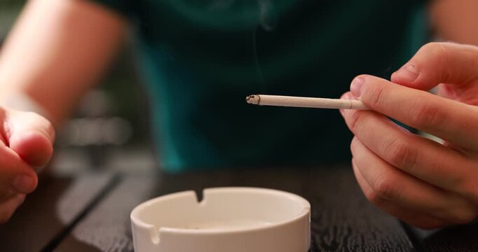 Man flicking ash from cigarette into ashtray at wooden table outdoors, closeup