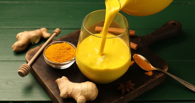 Golden milk turmeric latte. Pouring drink into glass at green wooden table, closeup