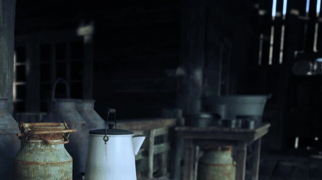 Inside an old barn, various milk containers are placed on wooden tables. Dim light comes through gaps, highlighting the rustic scene. The atmosphere shows a past farming life.