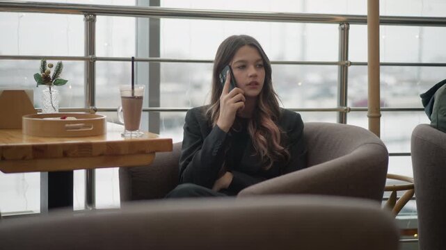 Young woman sits in curved upholstered cafe booth holding smartphone to ear, glancing sideways with attentive pensive expression while relaxed daylight fills modern urban interior, with woman, female