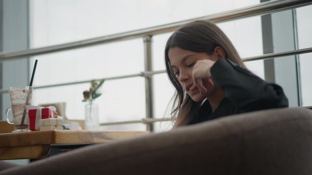 Teenage girl seated in cafe booth rests head on hand with eyes downcast, showing subtle downward movement into thoughtful calm under soft daylight from window, with quiet indoor mood and casual