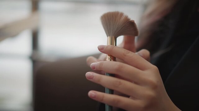 Close-up view of hands holding and aligning multiple makeup brushes with gentle fingertip movement, bristles facing upward beside bright window light, creating soft beauty prep and careful cosmetic