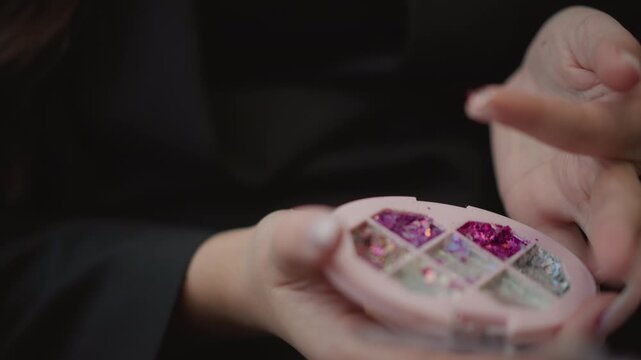 Close view of hands inside clear plastic container filled with pink and purple glitter and sequins, showing delicate tapping, picking, and sorting motions with soft reflective sparkle