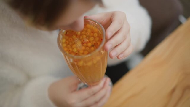Overhead close-up shows child holding transparent cup filled with glossy orange tapioca pearls, cup tilted upward while drinking, small hands gripping firmly and drink shifting gently