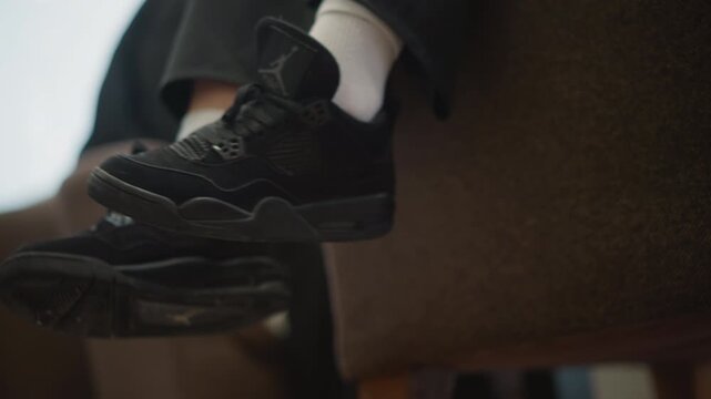Close-up low-angle view of child lower legs and feet in black sneakers and white socks dangling from upholstered chair, with subtle swinging movement and quiet indoor waiting mood, with child