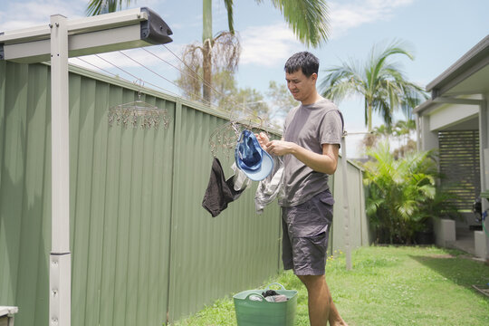 Man laundry hanging backyard housework chores domestic lifestyle, middle-aged Asian man drying clothes on a outdoor clothesline in a sunny suburban garden under a blue sky.