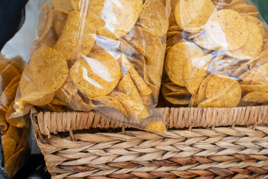 A square wicker basket filled with small, clear plastic bags of round, yellow organic corn chips.The homemade fried Mexican tortilla chips are packaged for sale at a market. The crispy chips are salty