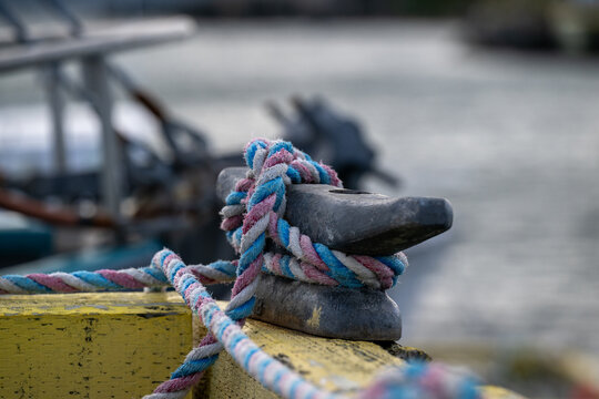 A metal mooring or bollard for boats on a yellow wooden wharf with a white, blue, and red braided fishing rope. The rope is tangled around the mooring base. There's harbour water in the background.