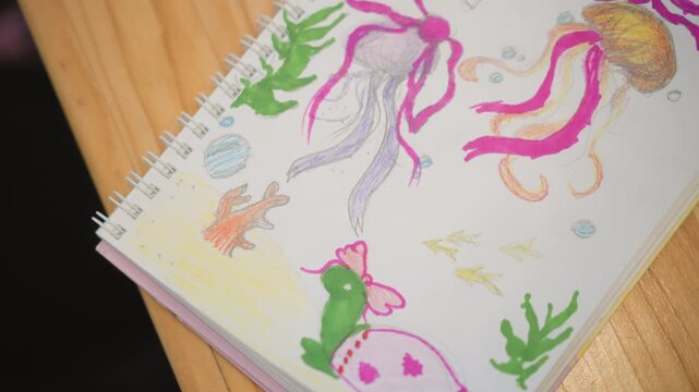 Angled close-up view shows sketchbook page being turned to reveal colorful underwater drawings with octopus tentacles, fish, coral, seaweed, and smiling whale across wood surface in soft indoor light