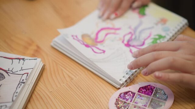Overhead close-up shows hands turning sketchbook pages over wooden table beside colorful nail palette and abstract drawings, with gentle browsing and careful creative review in soft natural light