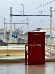 A bright red fire hydrant cabinet stands on a tiled elevated transit platform, with railway tracks and an approaching blue train visible in the background. © Anita Permatawaty