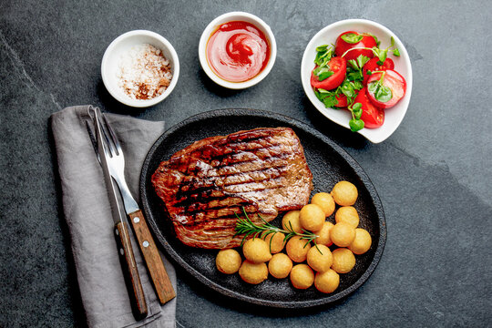 Grilled beef steak served on cast iron plate with tomato salad and potatoes balls. Barbecue, bbq meat beef tenderloin. Top view, slate background